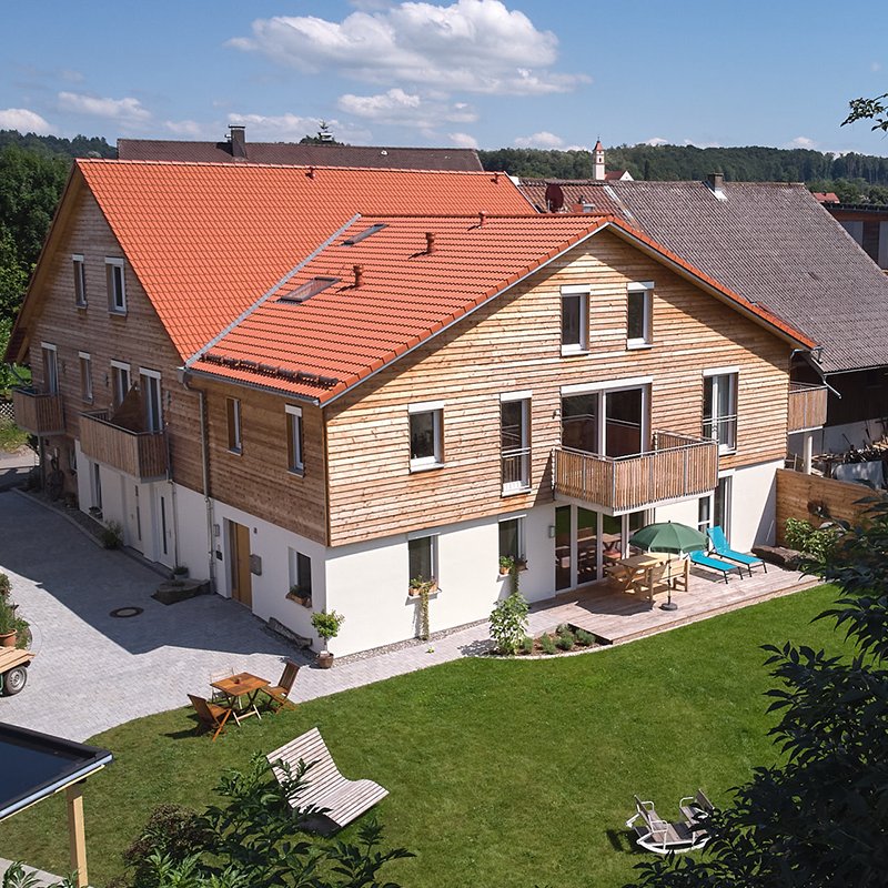 Rückansicht des Hauses mit zwei Balkonen, Holzverkleidung im Obergeschoss und heller Putzfassade im Erdgeschoss, umgeben von Rasen und Terrasse mit Gartenmöbeln.