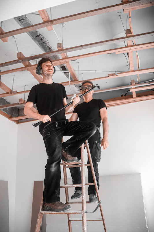Zwei Handwerker in schwarzer Arbeitskleidung bei der Deckenöffnung auf der Baustelle, einer steht lachend auf der Leiter mit Gehörschutz und Brecheisen, der andere sichert von unten.