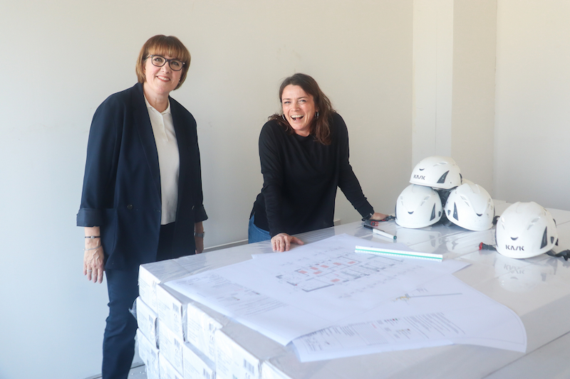 Zwei Frauen stehen an einem Tisch mit Bauplänen, mehreren weißen Schutzhelmen und einem Lineal. Beide lächeln – die Szene zeigt eine entspannte, professionelle Besprechung auf der Baustelle.