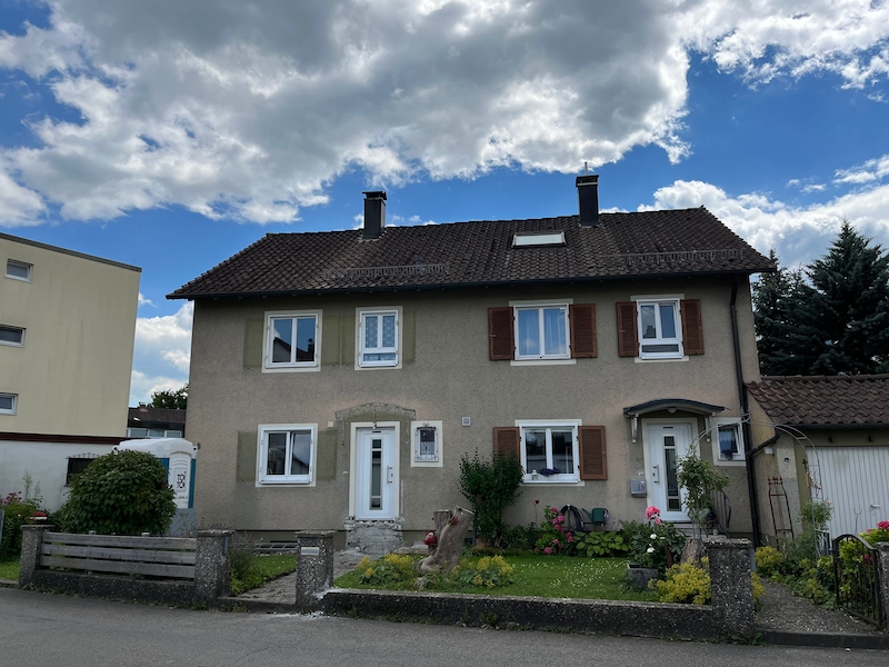 Frontalansicht einer Doppelhaushälfte mit Putzfassade, braunen Fensterläden und Satteldach. Der Zustand zeigt den sanierungsbedürftigen, aber gepflegten Altbestand mit Vorgarten und zwei Eingängen.