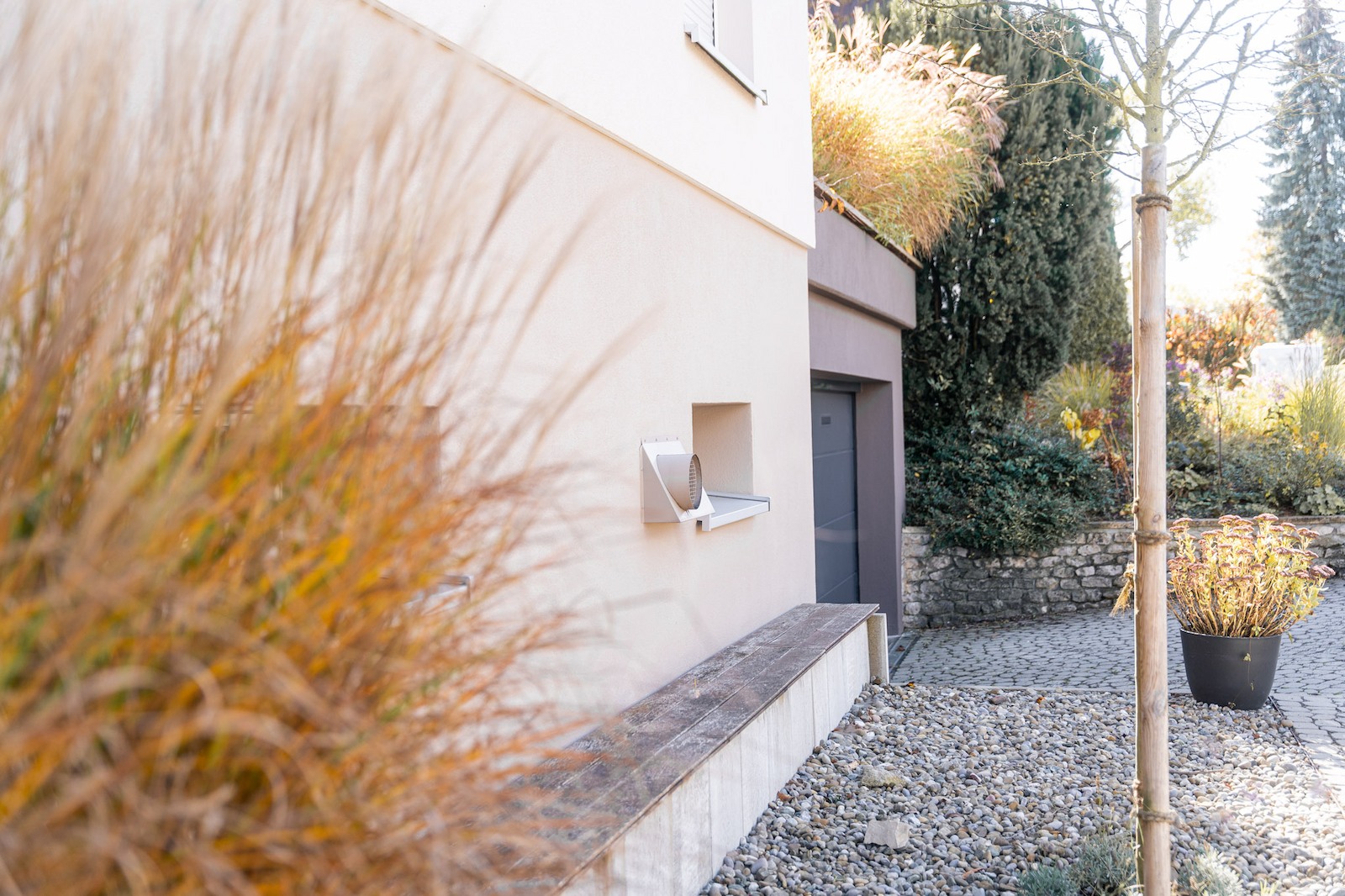 Außenansicht eines modernisierten Wohnhauses mit hellem Putz, integrierter Sitzbank und bepflanztem Vorgarten; im Vordergrund Ziergräser und ein junger Baum auf Kiesfläche.