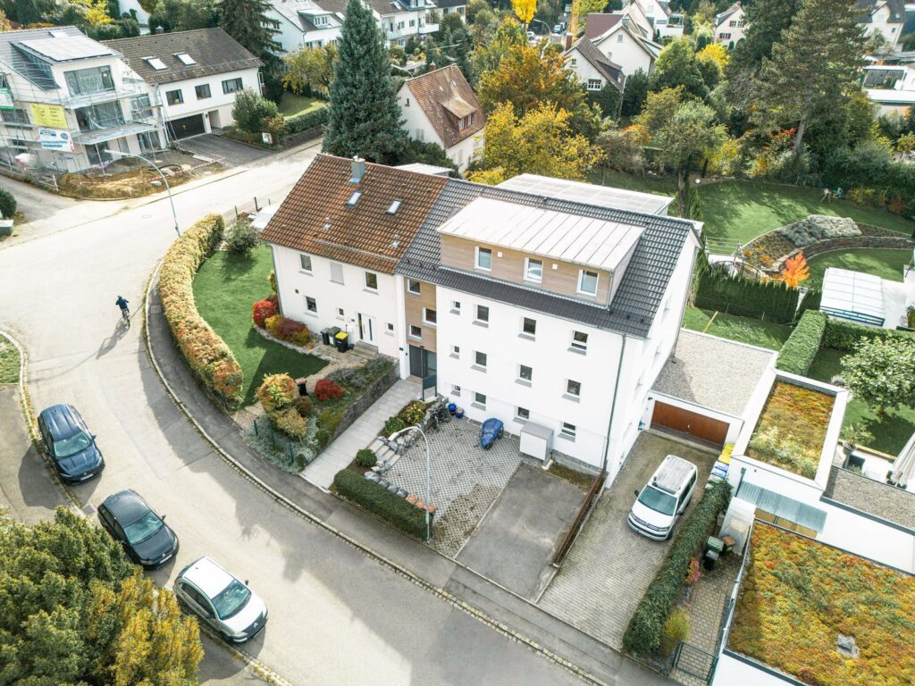 Luftaufnahme eines Eckgrundstücks mit saniertem Gebäude, sichtbarer Aufstockung, Vorgarten und zwei unterschiedlichen Dachformen. Die Umgebung zeigt gepflegte Gärten und gewachsene Nachbarschaftsstruktur.