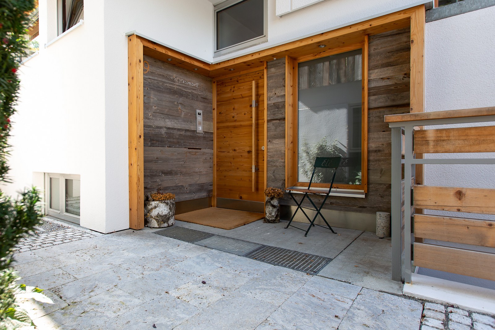 Eingangsbereich eines Wohnhauses mit einer Kombination aus weißer Putzfassade und Altholzverschalung. Der überdachte Eingangsbereich ist mit einem schlichten Stuhl, Pflanzgefäßen und Holztür gestaltet.