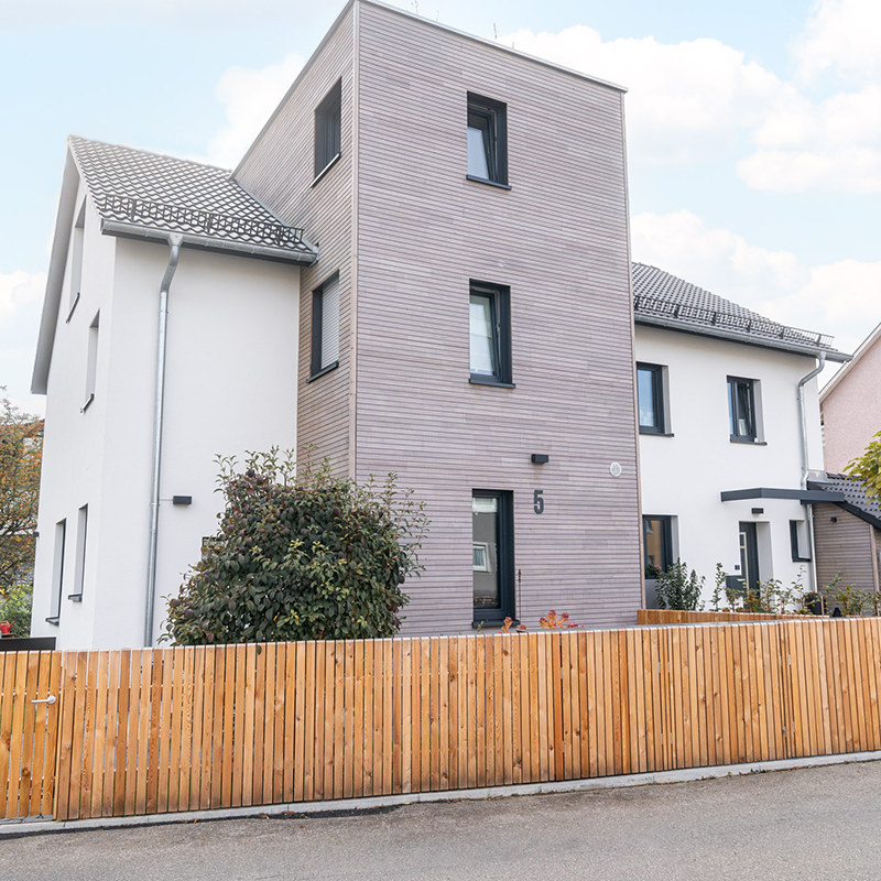 Das Wohnhaus von der Straße aus gesehen, mit hell verputztem Hauptbau, Holzfassade am Anbau und harmonischem Zaun.