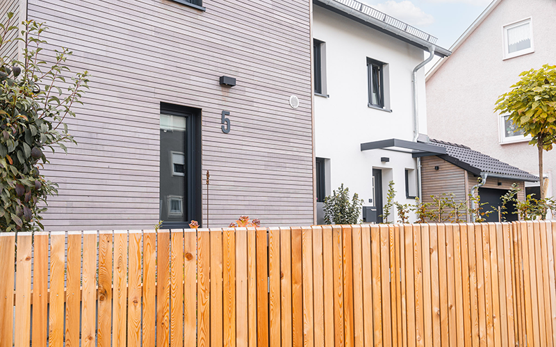 Das Wohnhaus von der Straße aus gesehen, mit hell verputztem Hauptbau, Holzfassade am Anbau und harmonischem Zaun.