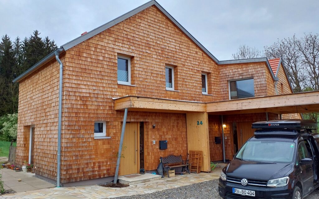 Frontansicht des Hauses mit traditioneller Holzschindelfassade, Satteldach, überdachtem Eingangsbereich und Carport. Vor dem Eingang befinden sich ein schwarzer VW Caddy, eine Bank und verschiedene Pflanzen.