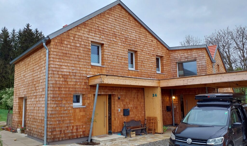Frontansicht des Hauses mit traditioneller Holzschindelfassade, Satteldach, überdachtem Eingangsbereich und Carport. Vor dem Eingang befinden sich ein schwarzer VW Caddy, eine Bank und verschiedene Pflanzen.