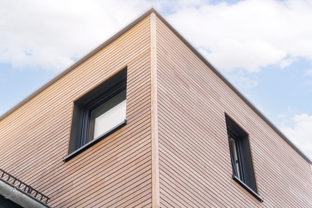 Nahaufnahme einer modernen Gebäudeecke mit horizontaler Holzfassade und zwei dunkel gerahmten Fenstern unter einem Flachdach, bei blauem Himmel.