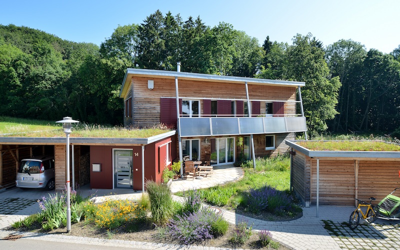 Modernes, zweigeschossiges Holzhaus mit begrünten Dächern, Solar­elementen und Carports. Vor dem Gebäude befinden sich Blumenbeete, Fahrrad und Auto – eingebettet in eine naturnahe Umgebung mit Bäumen.