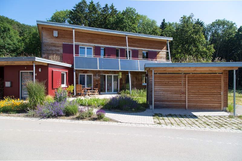 Modernes Wohnhaus aus Holz mit begrüntem Vorgarten, großem Balkon und Carport an sonnigem Tag.