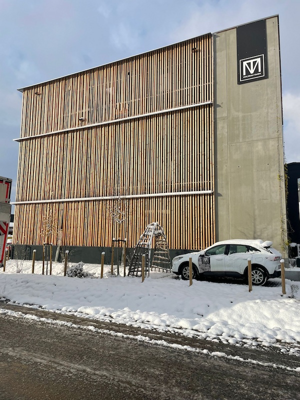 Moderne Gebäude­fassade aus vertikalen Holzlamellen neben einem geparkten Auto im Schnee.