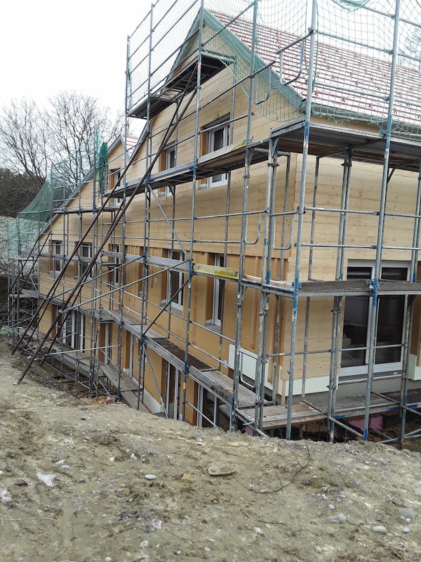 Mehrfamilienhaus im Holzbau mit Gerüst während der Bauphase, Fassade aus Holzplatten und Satteldach mit Ziegeln.